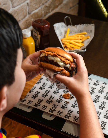 Cliente segurando um burger na Melt, com fritas na mesa e papel da casa.